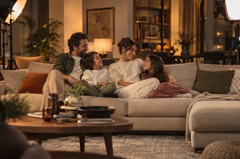 Dekore edilmiş salon setinde yeni bir kanepede dinlenen aile; görüntünün kenarlarında stüdyo ışıkları görünüyor.